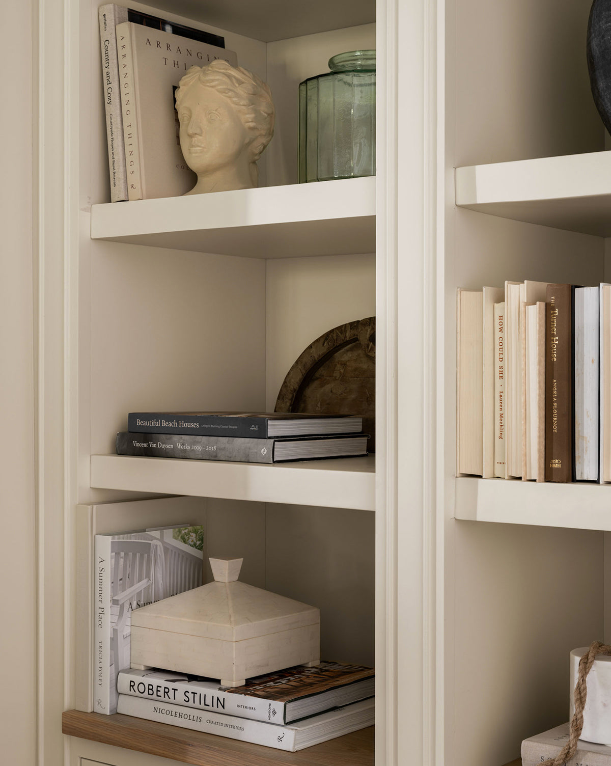 A beige bookshelf features neatly arranged books, the Hestia Bust in composite stone, a green glass jar, a decorative box, and a wooden tray for a minimalist, elegant look with artistic decor.
