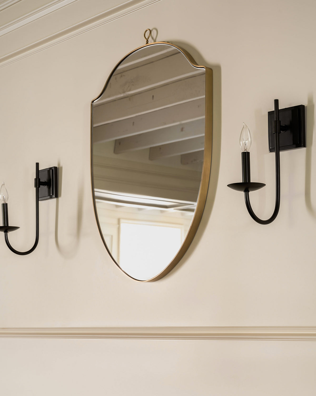 The Allegheny Wall Mirror, featuring a gold frame and curved edges, hangs on a beige wall between two black modern wall sconces with candle-style bulbs, reflecting a window and ceiling beams, by McGee & Co.