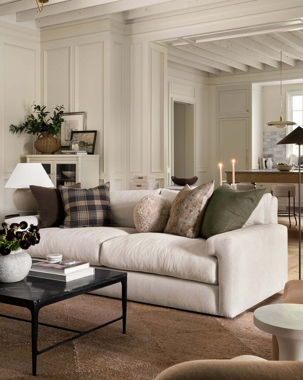 A cozy, modern living room showcases the Community Beckstead Sofa 92 with assorted throw pillows, a black coffee table, and candles. Neutral tones and layered textures create a warm, inviting vibe. The dining area appears in the background, featuring accents from McGee & Co.