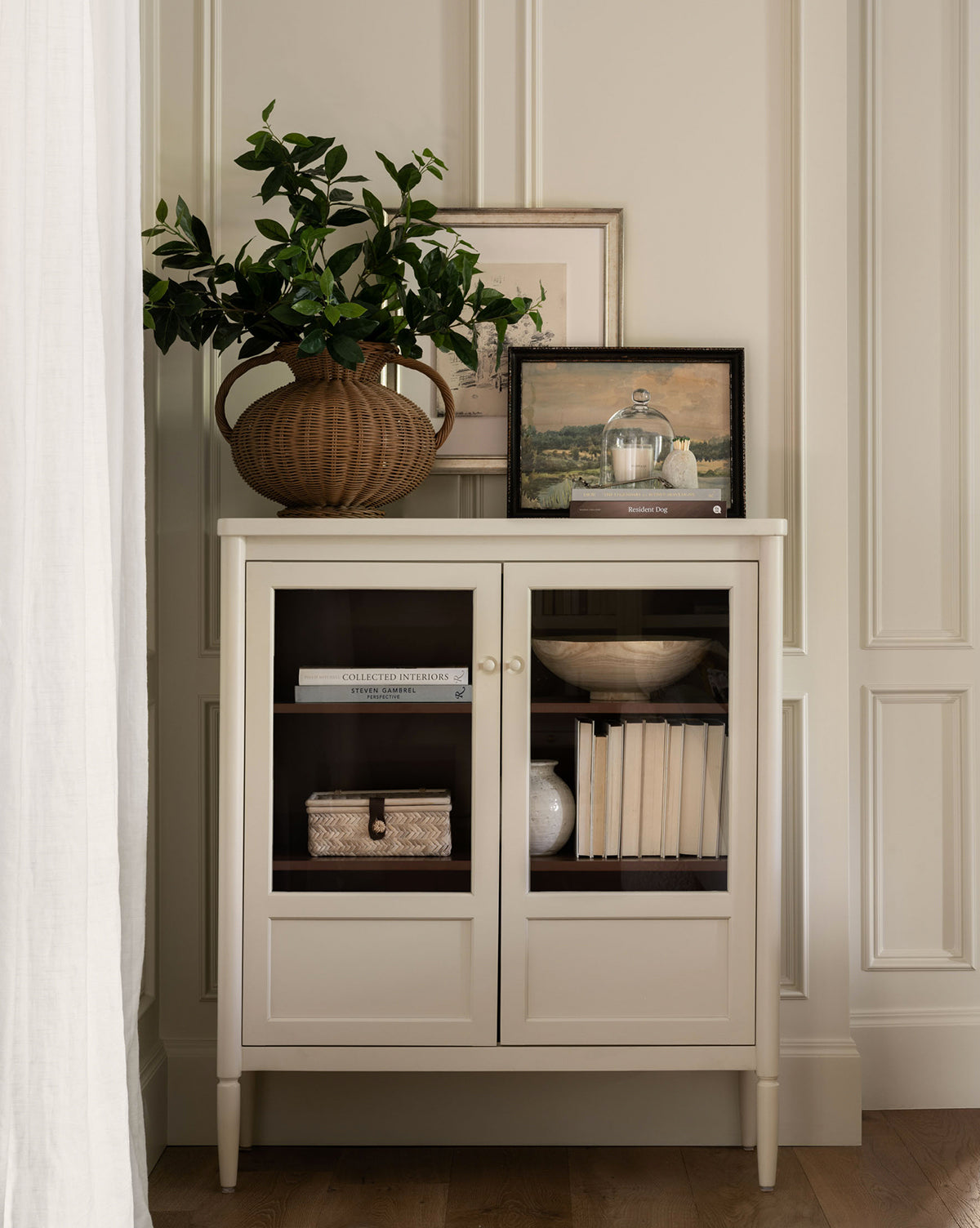 A bone white Highgate Cabinet with glass doors showcases books, a woven box, and decor. On top are framed art, a wicker vase with green branches, and more books. The scene is bright and elegant with Victorian-inspired charm, by McGee & Co.