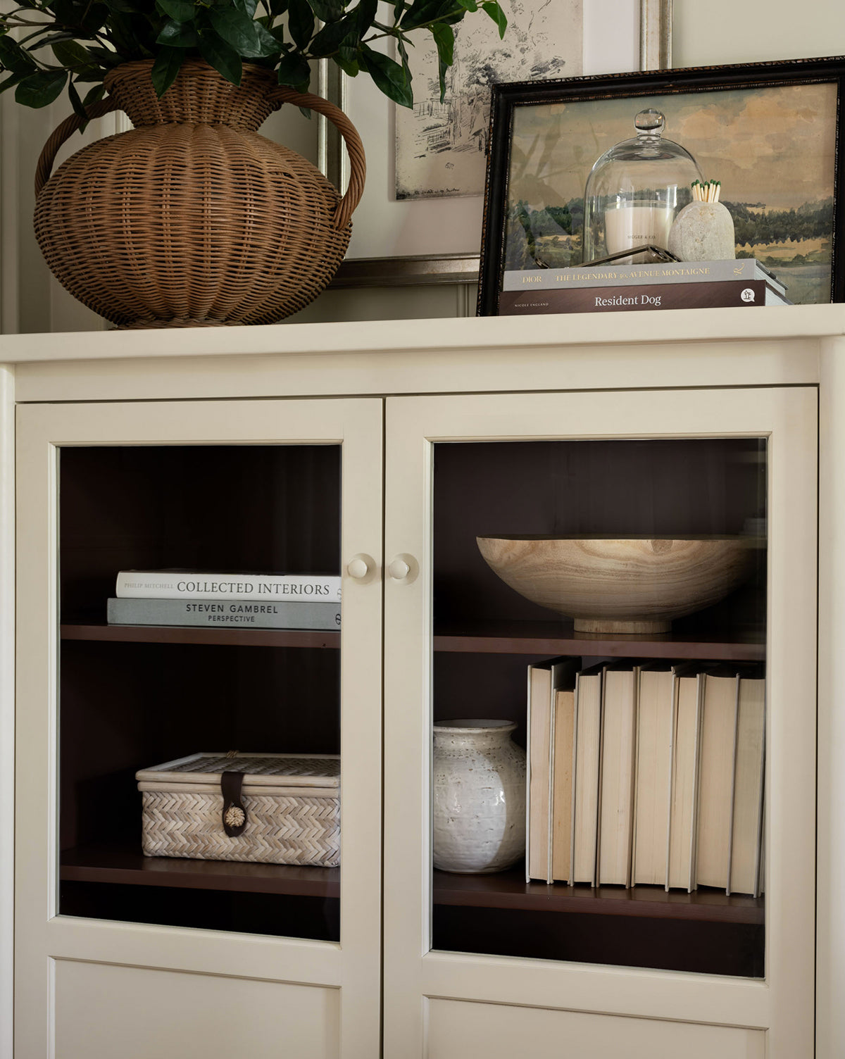 A cream-colored cabinet with glass doors displays books, a woven box, a ceramic vase, and a wooden bowl. On top sit a wicker jug with greenery, framed art, kitchen décor, and a Glass Cloche covering small items or baked treats.