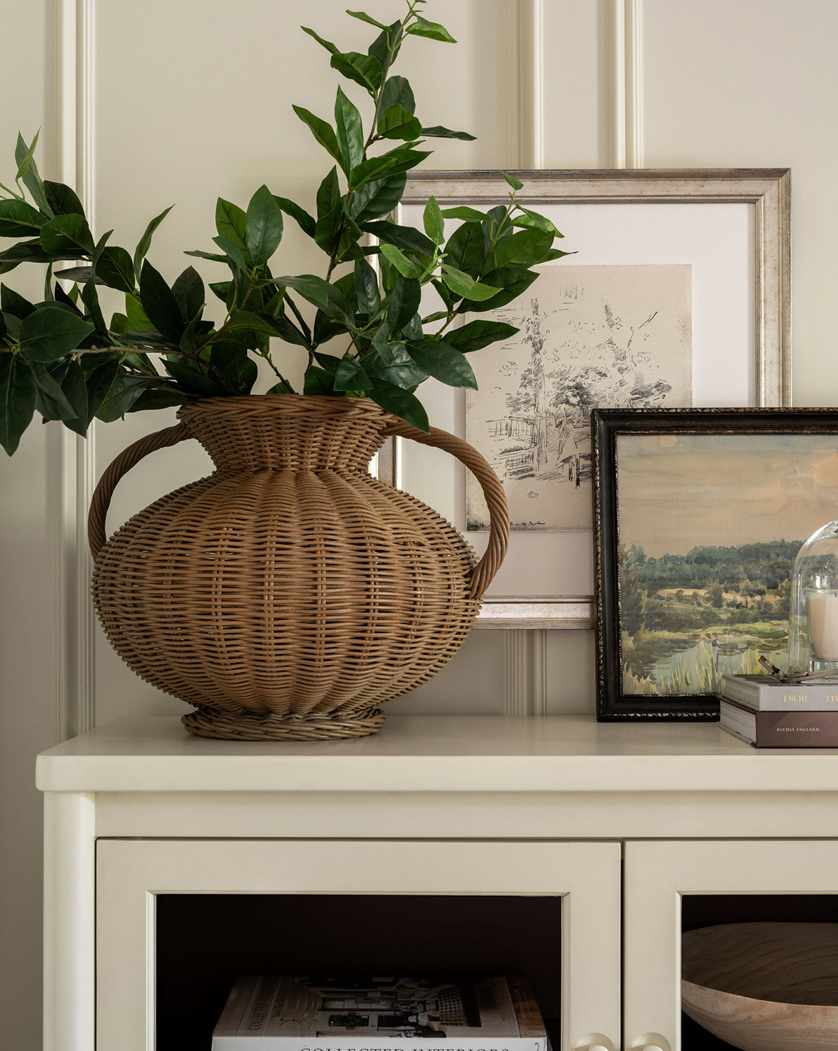 The Antonia Woven Vase with green leafy branches sits on a cream cabinet, beside two framed art prints, a stack of books, and a glass cloche holding a candle.