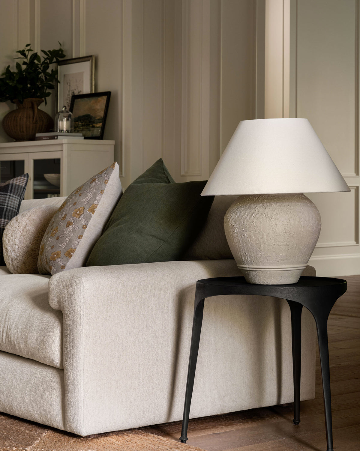 A cozy living room corner with a beige sofa and assorted pillows sits next to a black side table topped with the Abbott Ceramic Table Lamp, featuring a round ceramic base and white linen shade, creating a warm, softly lit atmosphere by McGee & Co.