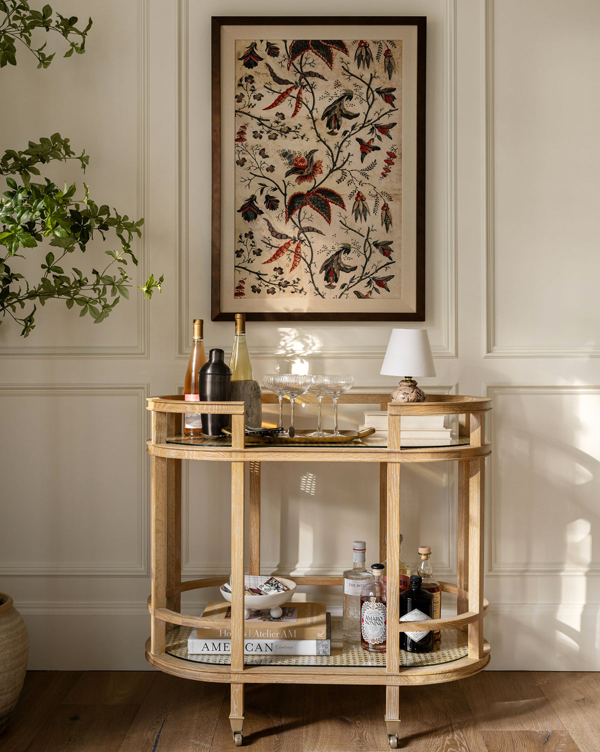 The Berries & Leaves bar cart holds bottles, glasses, and a lamp against a cream wall with vintage bronze-framed art. A leafy plant sits nearby; books, bowls, and more bottles fill the lower shelf.