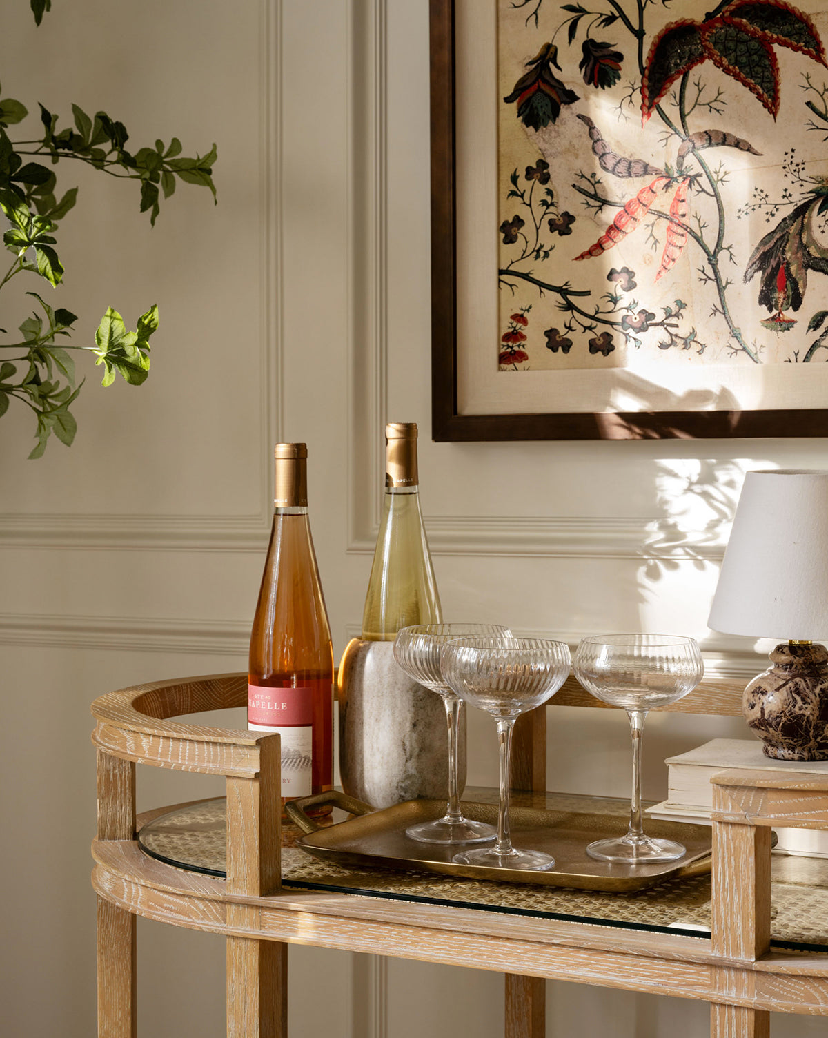 A wooden bar cart holds two wine bottles and three crystal glasses on the Adrienne Brass Tray beside a small lamp, with sunlight casting shadows on botanical art and green leaves peeking in from the edge.
