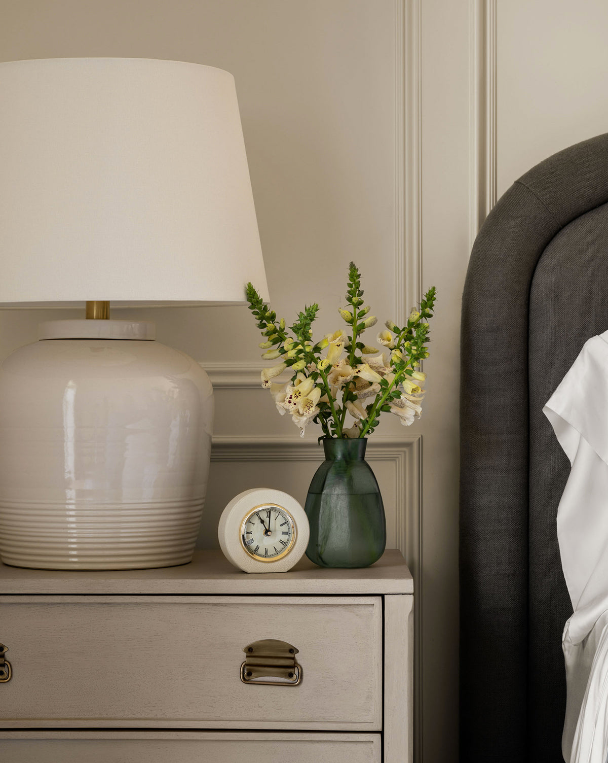 A bedside table displays a Shagreen Table Clock, a large white lamp, and a green vase with yellow and white flowers. The gray upholstered beds edge and white bedsheet appear on the right.