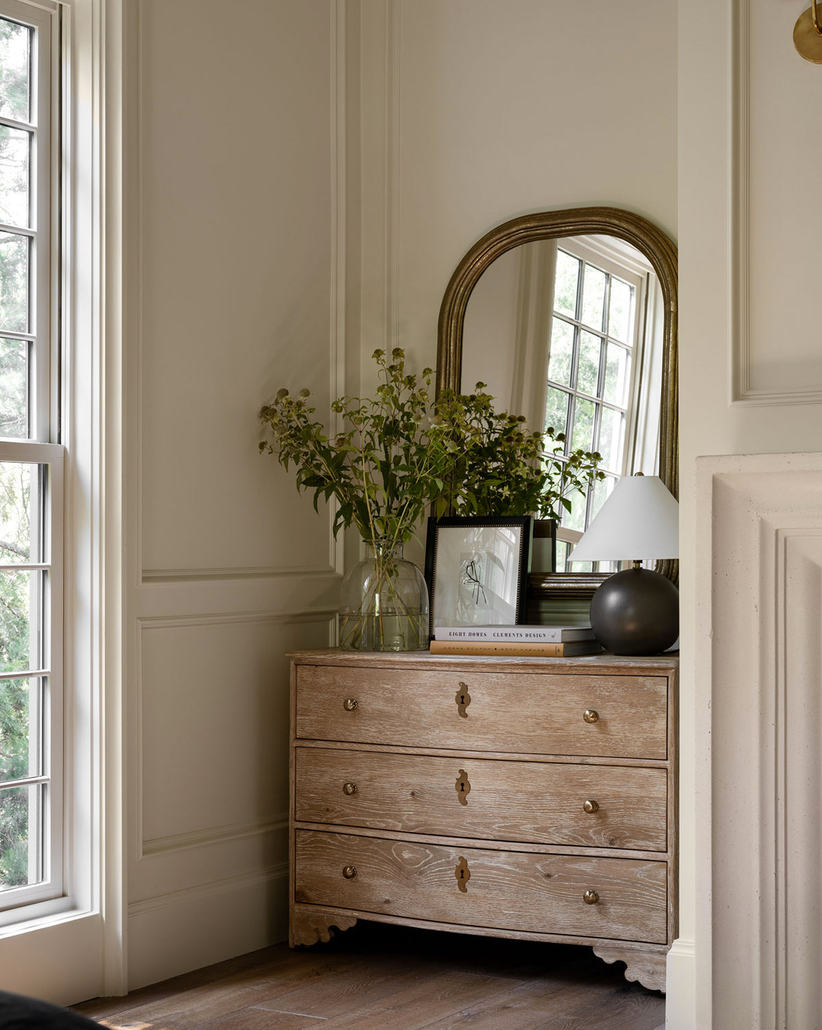 The Cybil Dresser, with brass handles, stands by a window. On top are a glass vase of greenery, framed art, books, a small white lamp, and a large arched mirror reflecting the window’s natural light by McGee & Co.