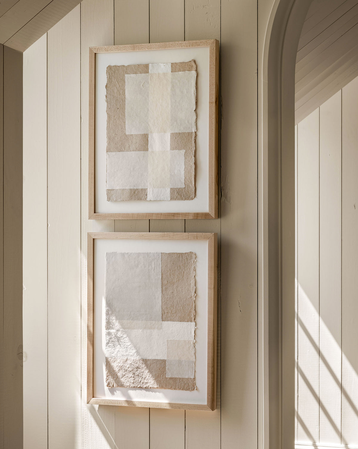 Collage 1, a framed abstract artwork crafted from handmade paper in neutral beige and white tones, hangs vertically on a light wood-paneled wall near an arched doorway. Sunlight casts gentle shadows across this neutral wall art.