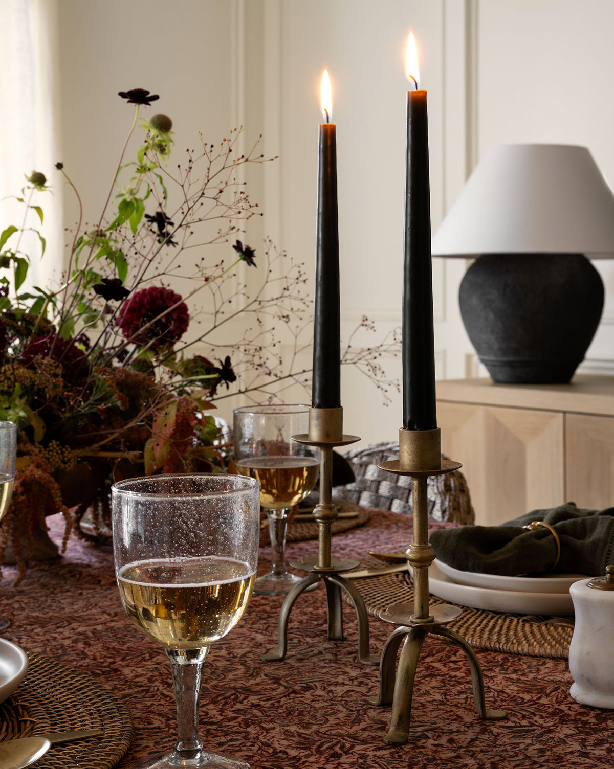An elegant dining table is set with glasses of white wine, a floral centerpiece, and two lit black candles in antique brass Claridge Taper Holders, creating a warm atmosphere. A lamp glows softly in the background.