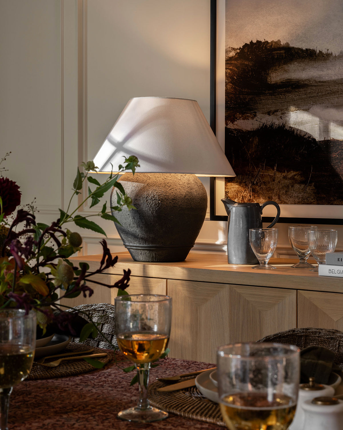 A cozy dining room features a rustic table set for a meal with golden-hued glasses, dark floral centerpiece, and the Abbott Ceramic Table Lamp with a white linen shade and round ceramic base on the wooden sideboard among art and glassware, by McGee & Co.