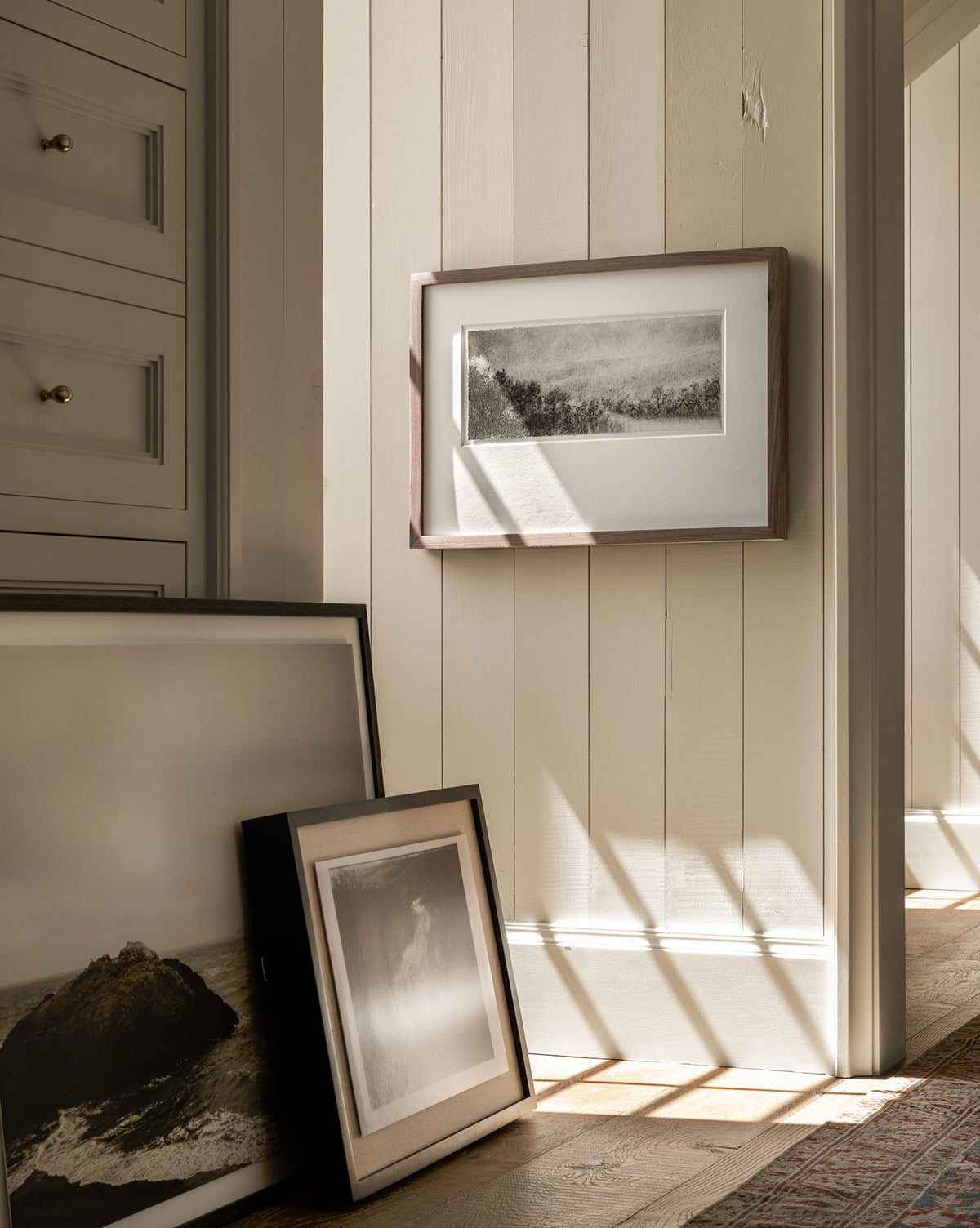 Hill Country Study I by Aileen Fitzgerald, a framed black-and-white artwork, leans against a beige paneled wall with sunlight casting shadows on the floor. One piece hangs above, and a dresser with brass knobs is partly seen on the left.