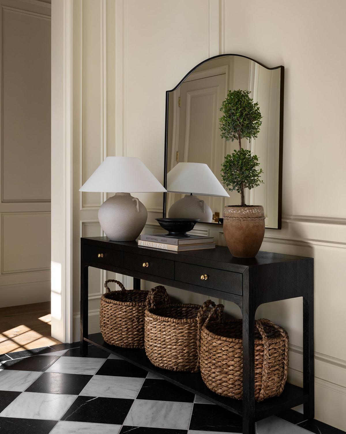 A black console table with three woven baskets beneath is styled with a Mays Table Lamp, a potted topiary, and a black bowl. A mirror hangs above, while classic black and white checkered tiles cover the floor by McGee & Co.