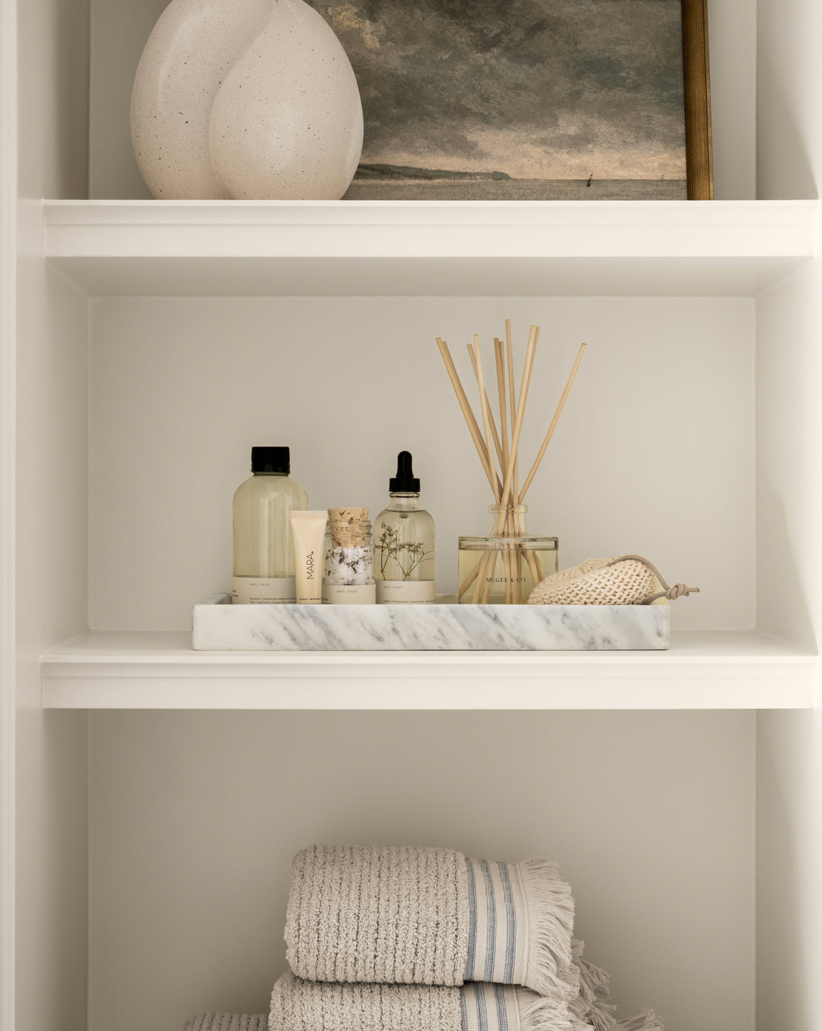 A Marble Tray displayed on white shelves holds a round vase, framed painting, reed diffuser, dropper bottle, and striped towels, creating a calm, spa-like decorative vignette.