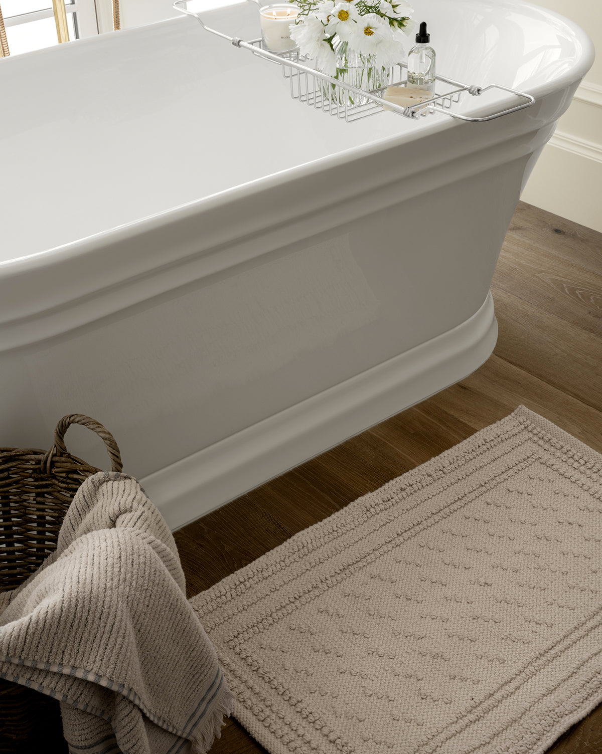 The Lohmann Bath Mat, in beige textured cotton, sits beside a white bathtub with a metal caddy and a wicker basket with a folded towel, creating cozy charm for your bathroom décor on the wooden floor.