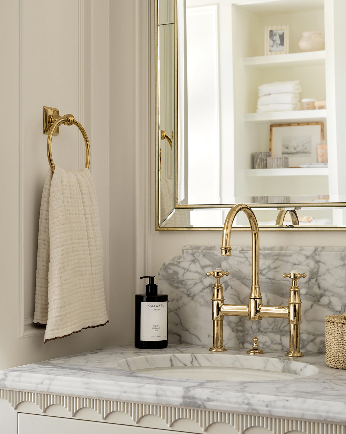 A bathroom sink with a marble countertop, gold faucet, and a gold towel ring holding a Cassidy Waffle Hand Towel. A soap dispenser is next to the sink, and shelves with towels and decor appear in the mirrors reflection.