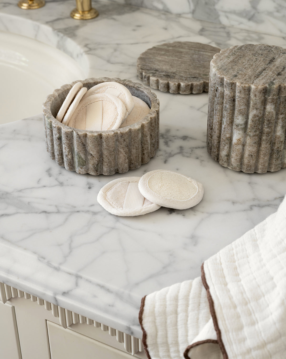 Round reusable cotton pads fill the Forster Marble Canister on a marble bathroom countertop, beside a cream towel with a brown edge—an elegant decor accent with organic materials by the sink.