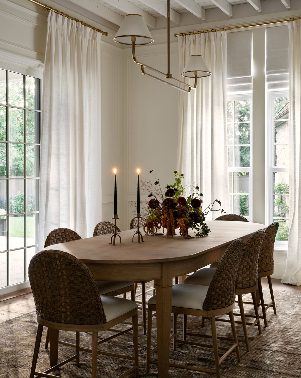 A dining room with a Hughes Oak Extension Dining Table, six woven chairs, two lit black candles, and a floral centerpiece. Large windows with sheer white curtains let in natural light, and a modern light fixture hangs above the table, by McGee & Co.