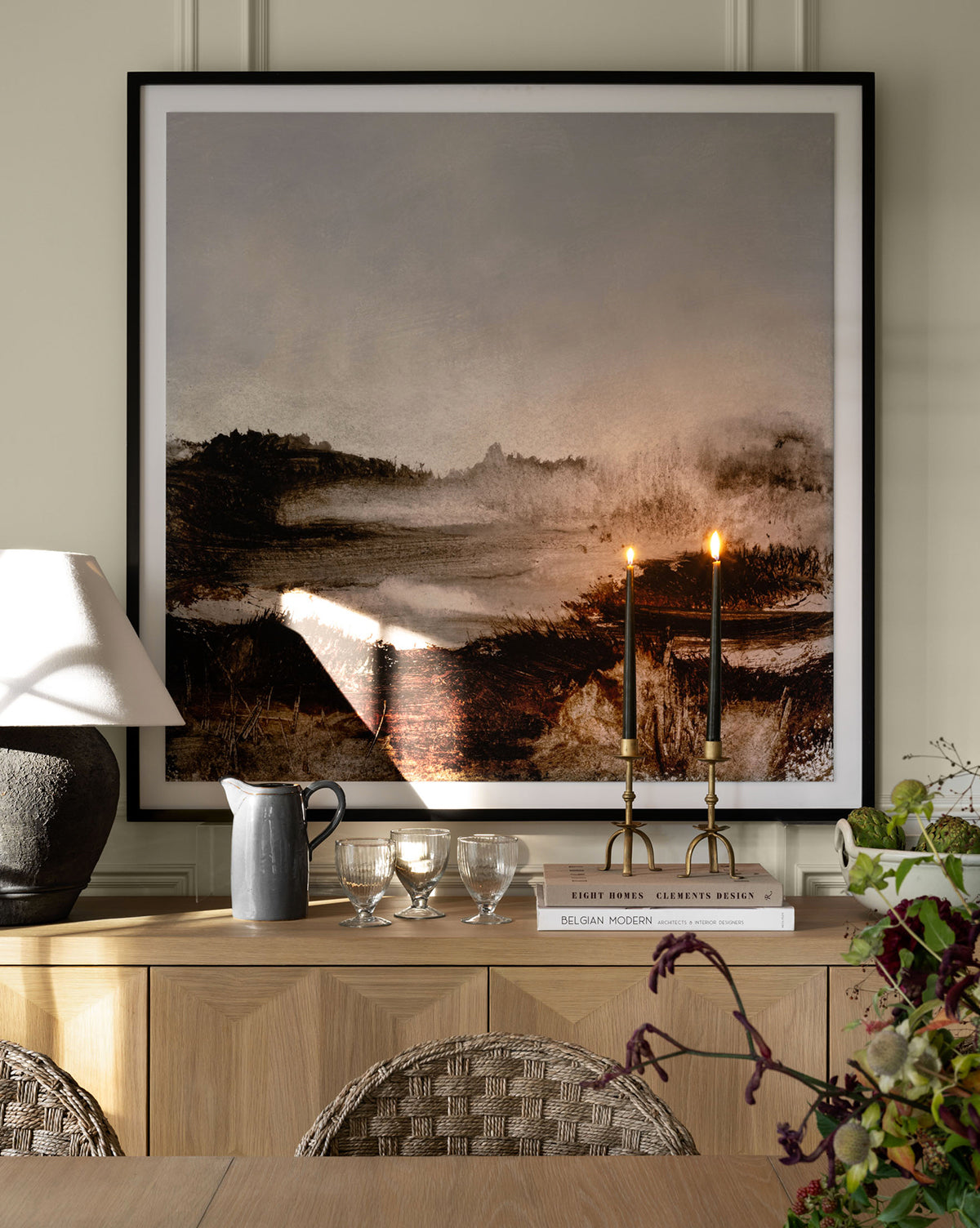 A wooden sideboard with decor sits below Woodbury by Dan Hobday, a large framed expressive landscape. Wicker chairs and a vase of flowers are partially visible in the foreground.