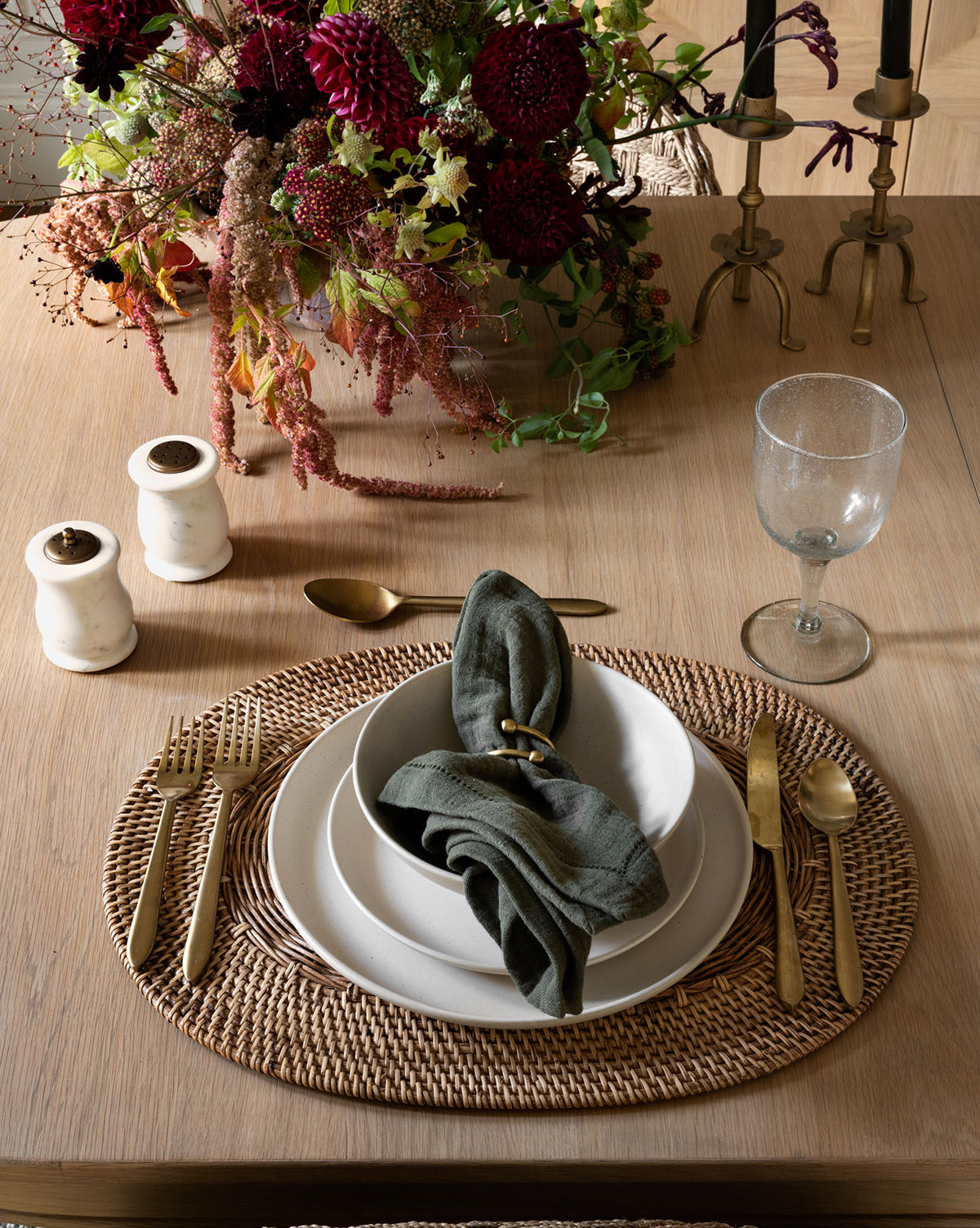 A wooden table set with a woven placemat, stacked white plates, a green cloth napkin, the Lilia Flatware (Set of 5), a wine glass, salt and pepper shakers, brass candlesticks, and a floral centerpiece with dark red flowers.