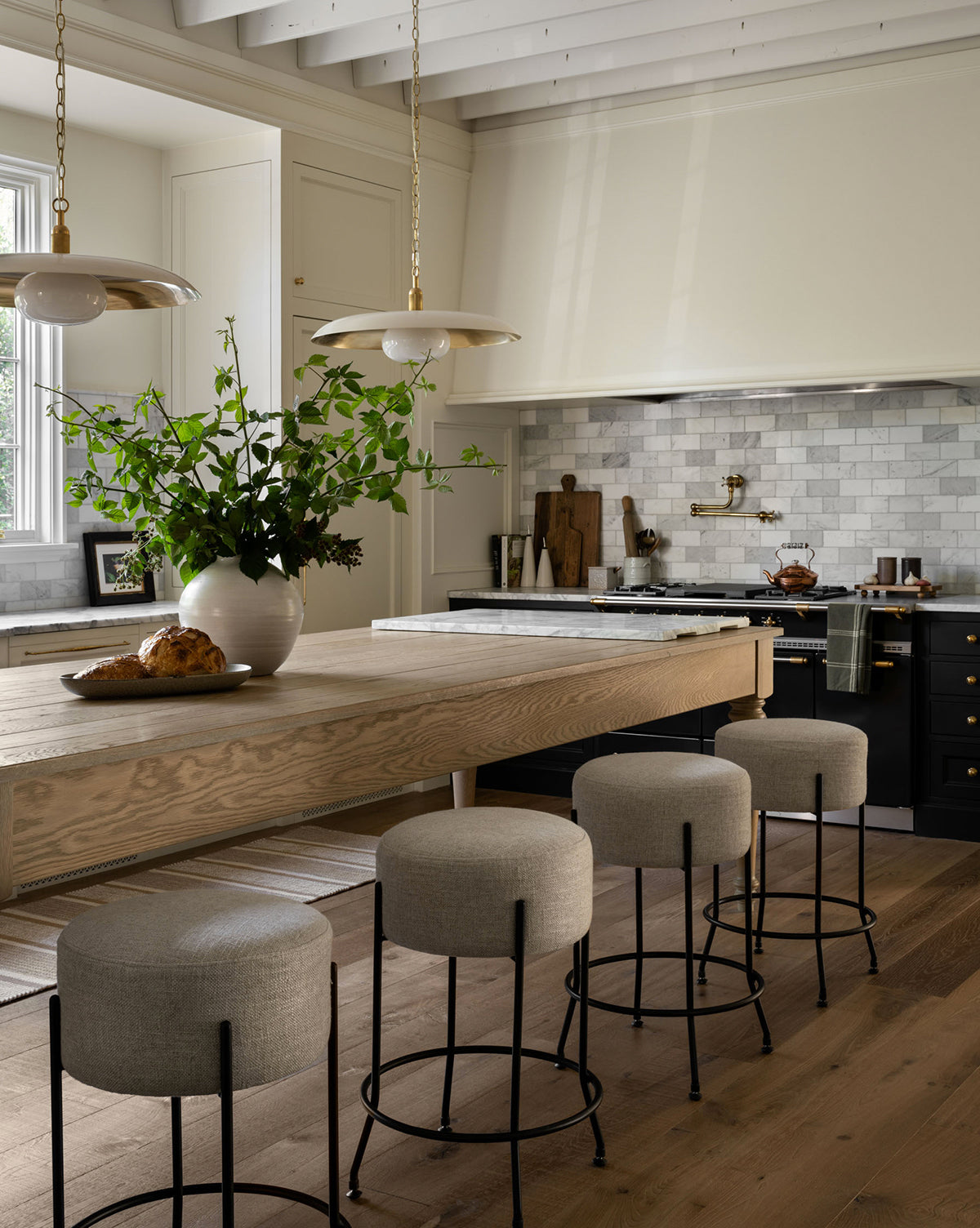 A modern kitchen features a wooden island, four gray upholstered stools, pendant lights, marble backsplash, black cabinetry with gold hardware, and the Enola Vase with green branches. Fresh bread decorates the island for a chic touch.