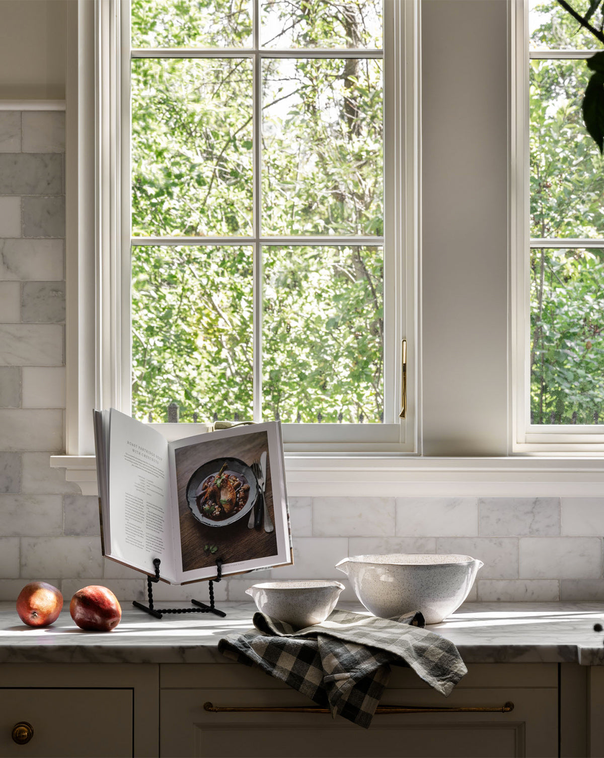A kitchen countertop styled with a cookbook on the Finlay Cookbook Holder, two peaches, a mixing bowl, a smaller bowl, and a checkered towel. Sunlight streams through a large window with green trees outside—perfect kitchen vibes.