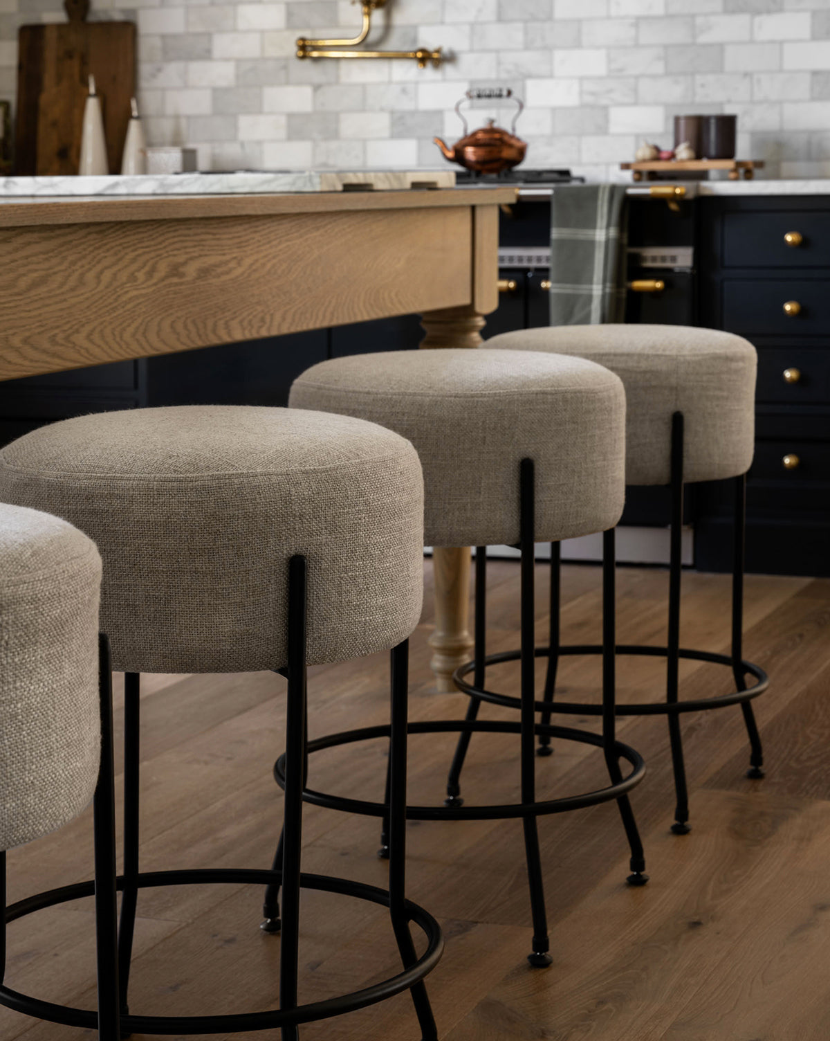 Three Makerspalm Evander Counter Stools with black metal legs are lined up at a wooden kitchen island, complementing a modern space with tile backsplash, dark cabinetry featuring brass hardware, and wooden flooring, by McGee & Co.