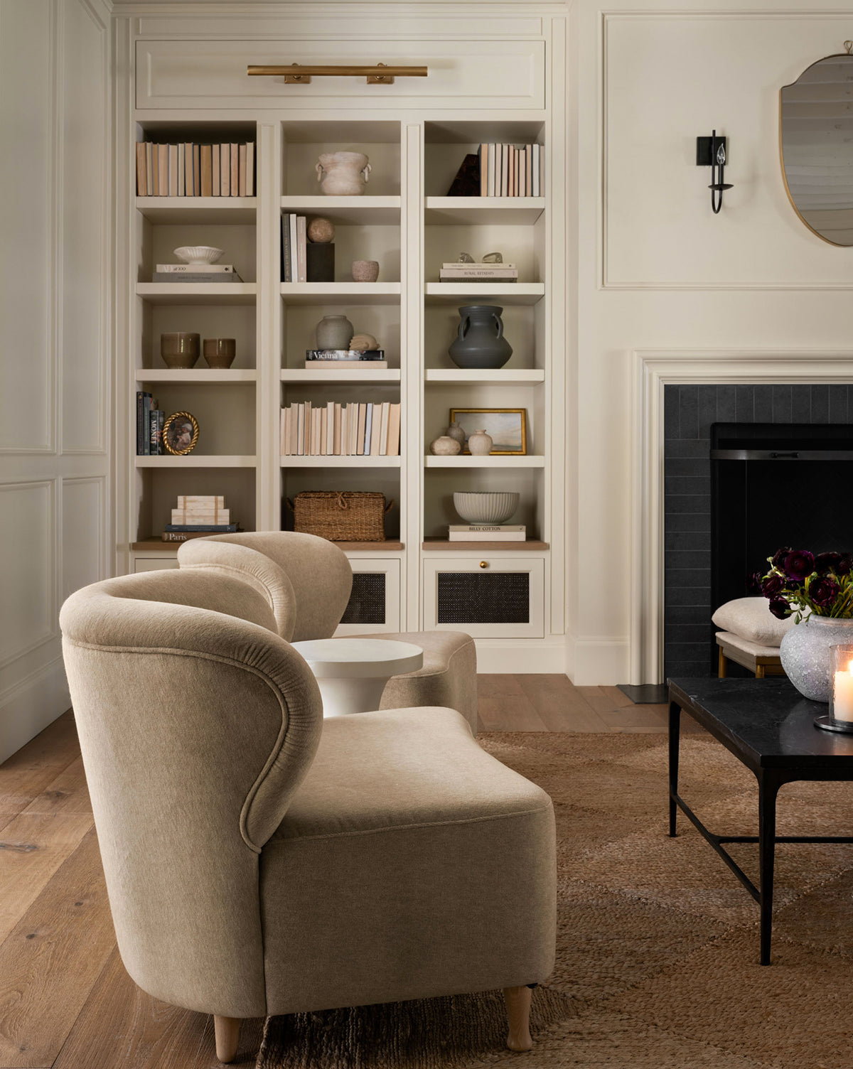 Two Carmella Slipper Chairs (Ready to Ship) in beige face a black coffee table in a cozy living room featuring a built-in bookshelf, dark fireplace, neutral rugs, and soft lighting for a warm, inviting atmosphere, by McGee & Co.