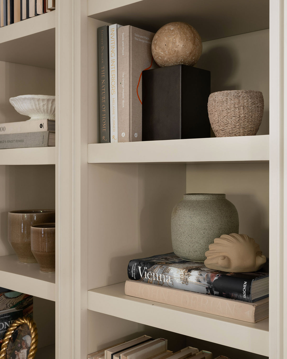 Beige shelves display stacked books, ceramic mugs, a ribbed bowl, a round stone accent, a woven basket, a large textured vase, and the Baroque Shell Object. Decor is minimalist and neutral-toned.
