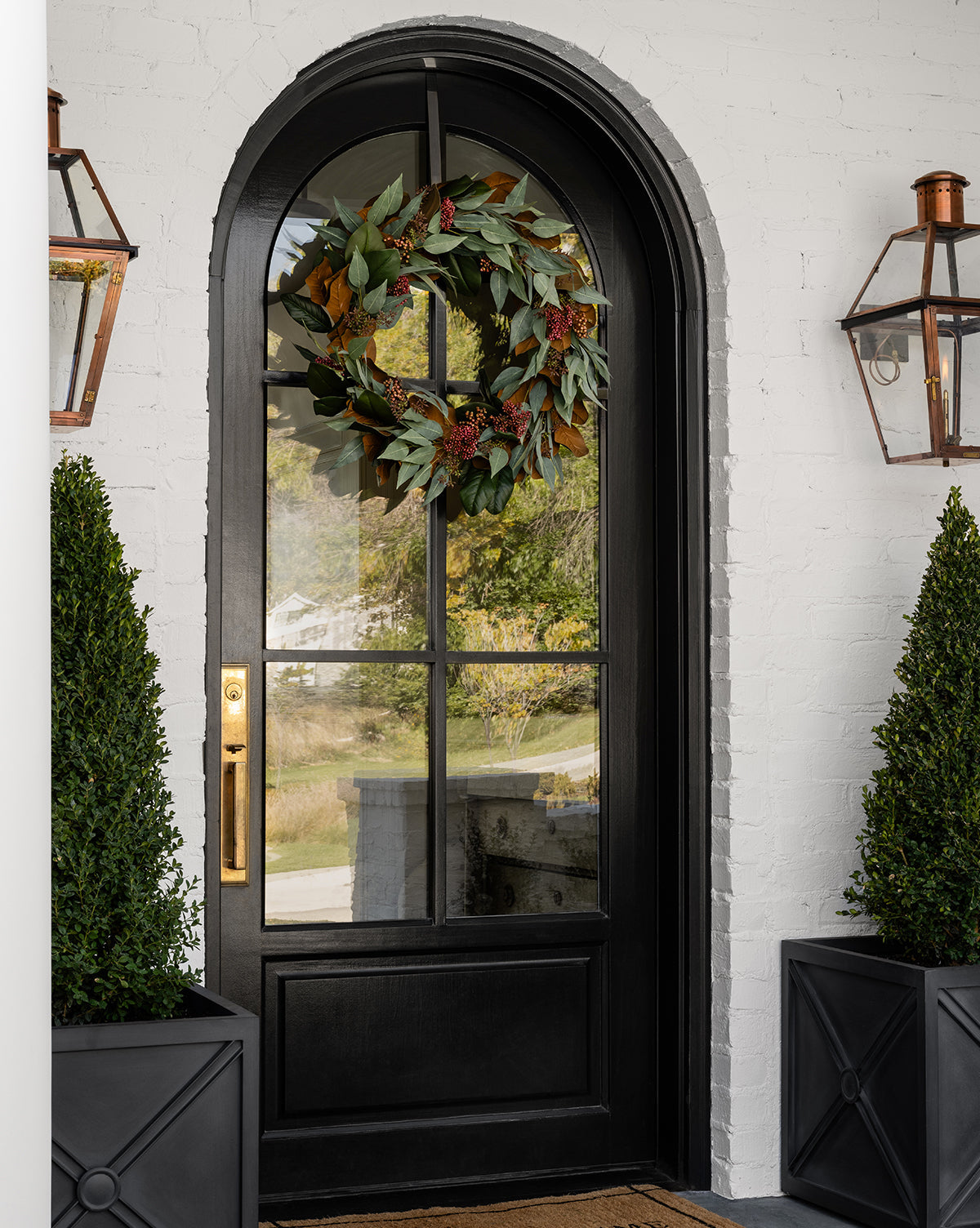 A black arched front door with glass panes and a brass handle features a lush 30 Faux Magnolia & Eucalyptus Wreath. Copper lanterns and large potted shrubs flank the entrance, set against a white brick wall for an elegant look.