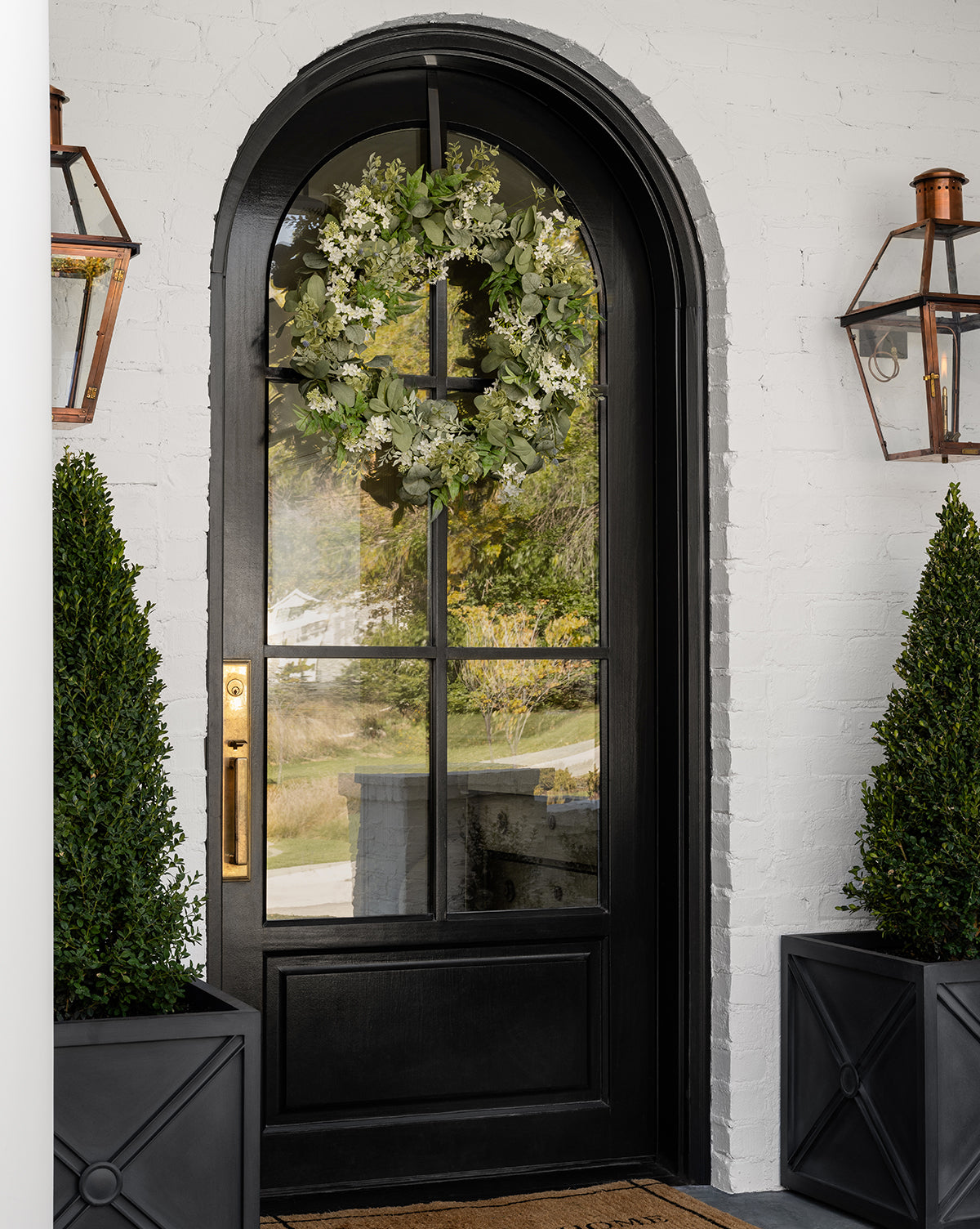 A black arched front door with glass panels displays an Orange Jasmine & Eucalyptus Wreath. The door sits in a white brick wall, flanked by two large planters with trimmed shrubs and copper lanterns on either side.