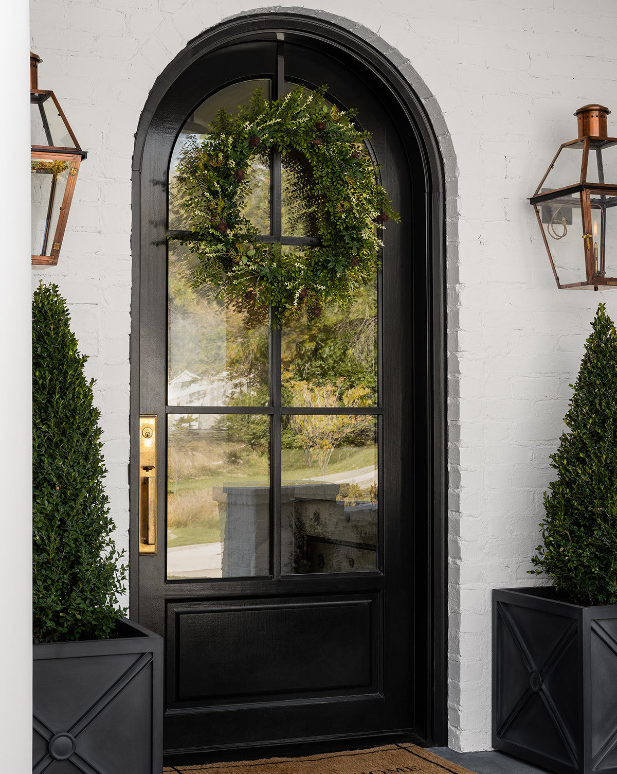 A black arched front door with glass panels, adorned with the Faux Summerset Wreath, flanked by potted boxwood plants and wall-mounted lanterns on a white brick exterior—perfect for seasonal decor.