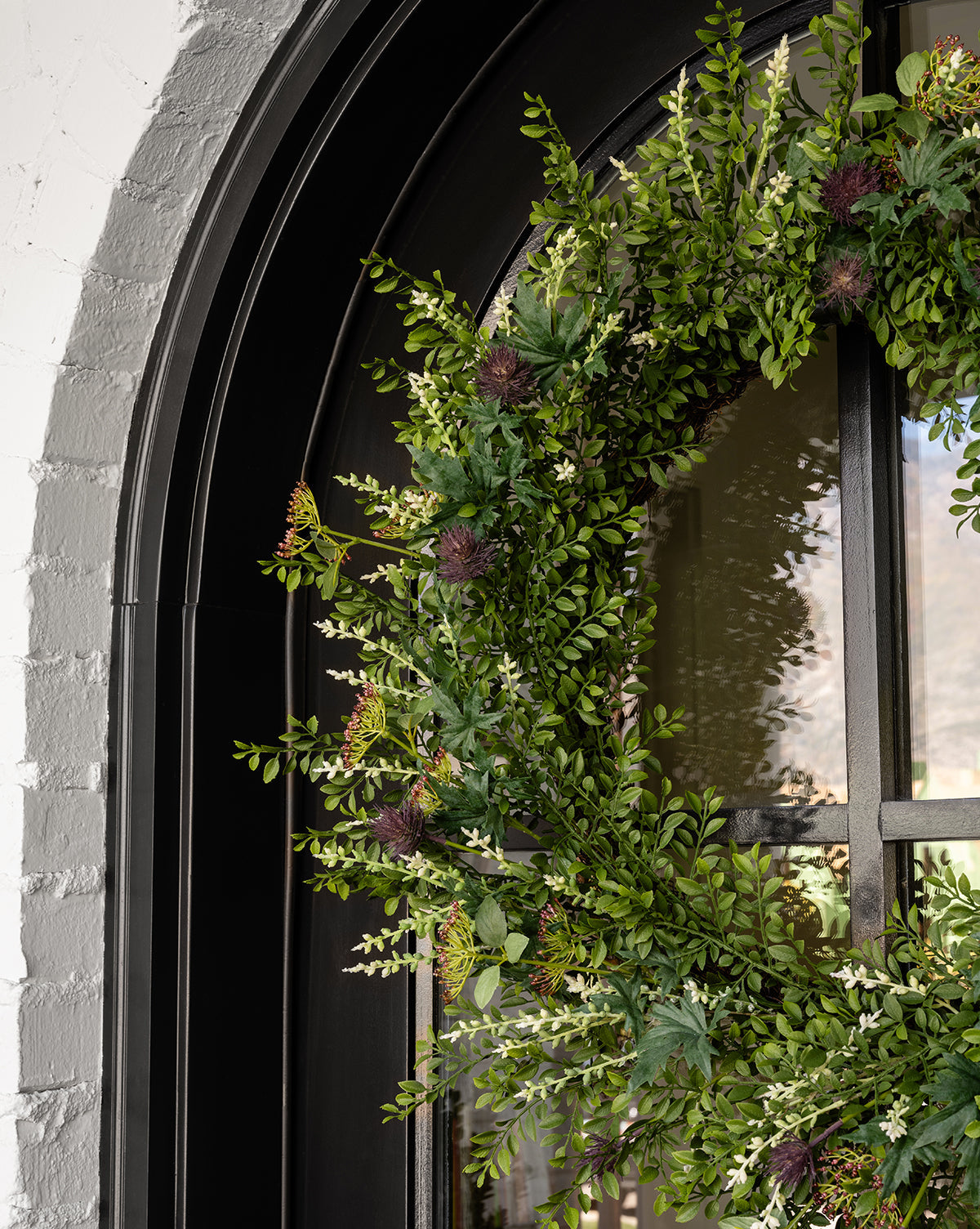 The Faux Summerset Wreath features lifelike leafy branches and flowers, hanging on a black arched door against a white brick wall—the perfect seasonal decor made from realistic faux greenery.