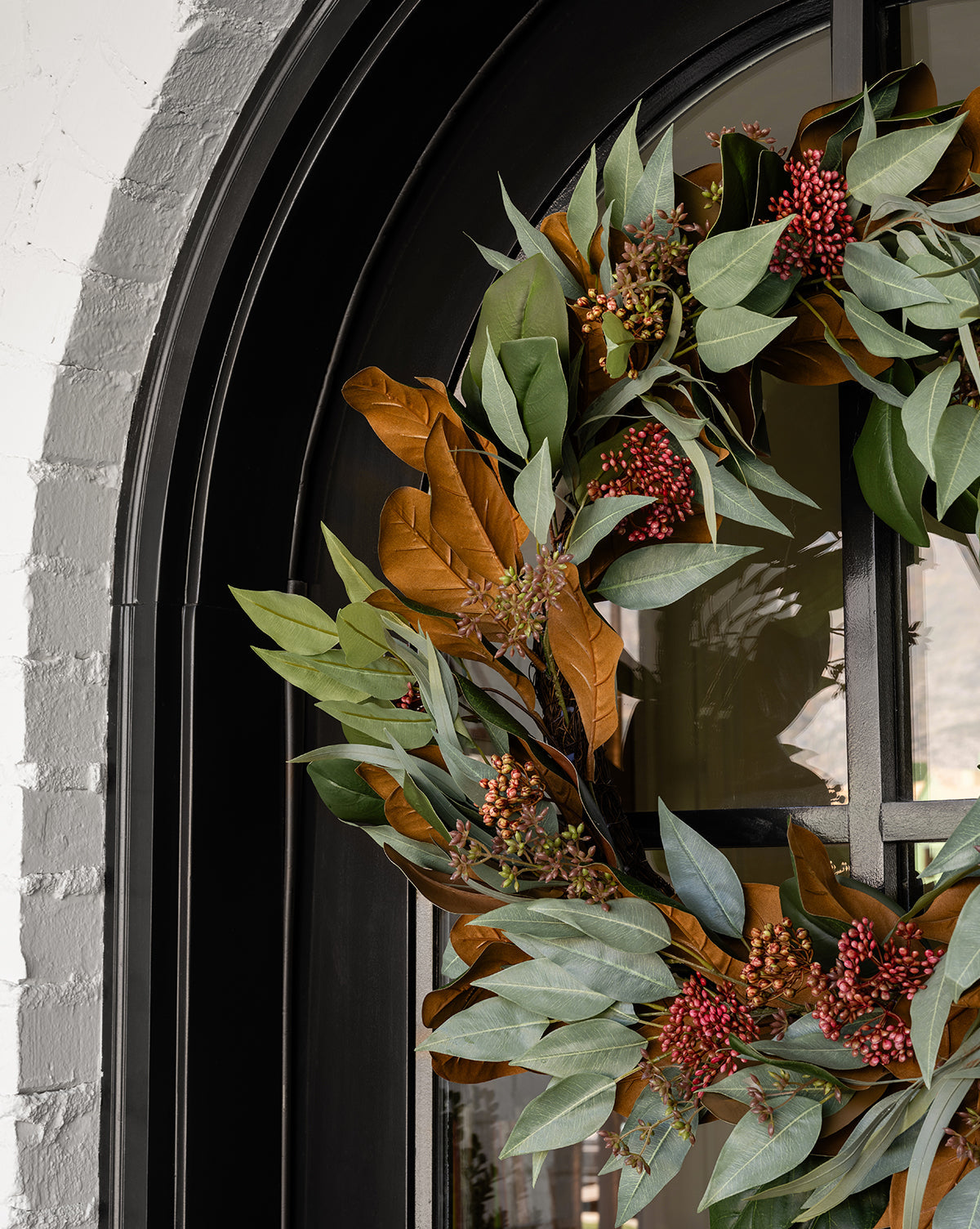 A 30 Faux Magnolia & Eucalyptus Wreath with green and brown leaves, red berry clusters, and magnolia accents hangs on a black arched door beside a white brick wall.