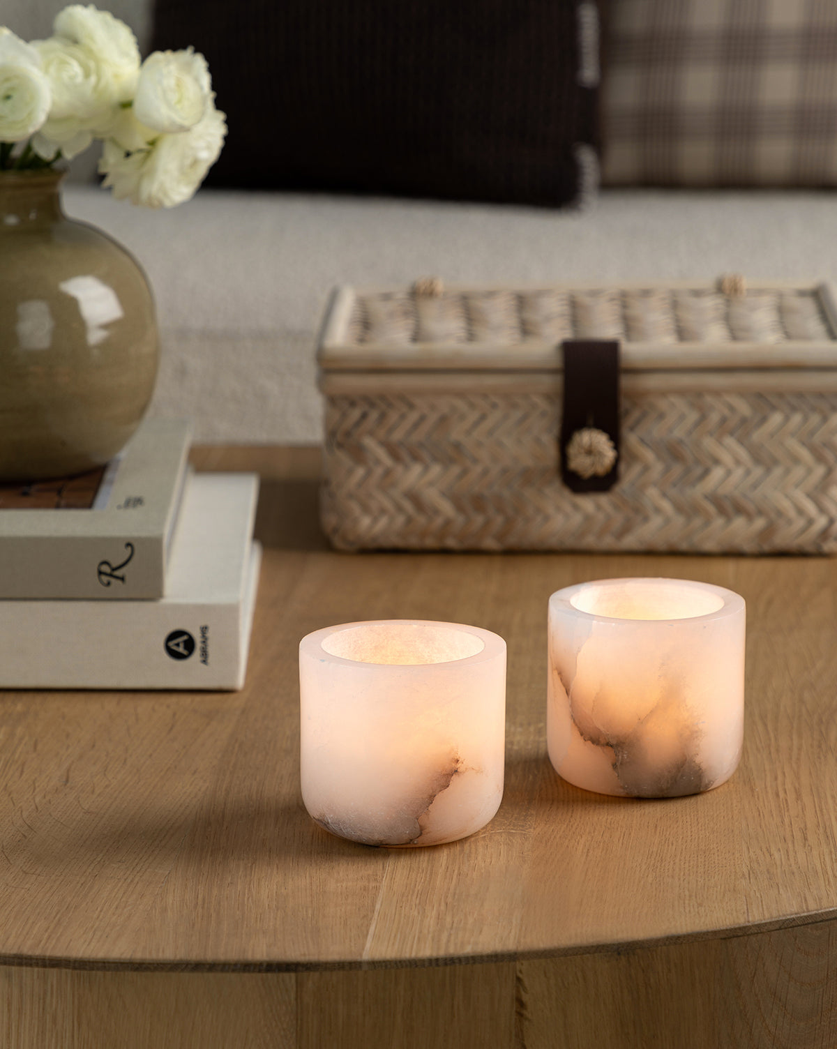 Two Alabaster Tea-light Holders with lit candles sit on a wooden coffee table, accompanied by stacked books, a vase of white flowers, and a woven basket in a cozy living room.