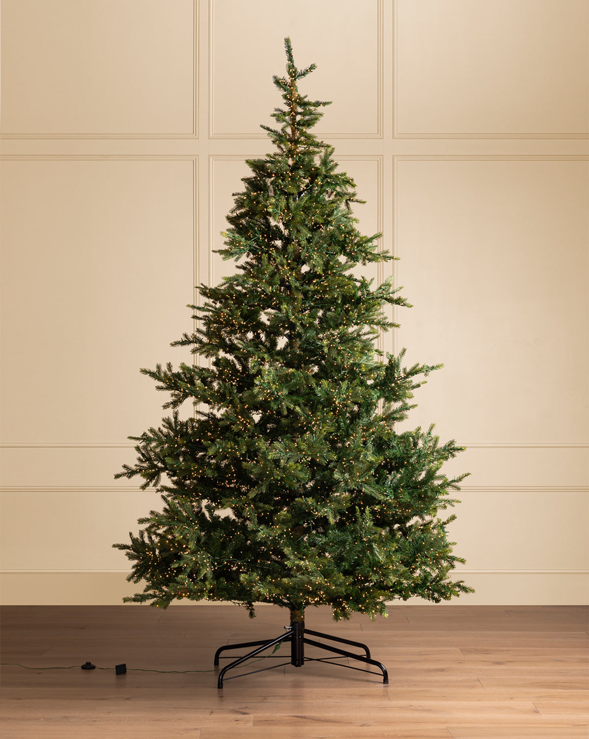 A 10' Grandis Fir Pre-Lit Faux Tree stands on a wooden floor by a cream paneled wall, its LED lights twinkling across dense branches, with a power cord trailing to the left, by McGee & Co.