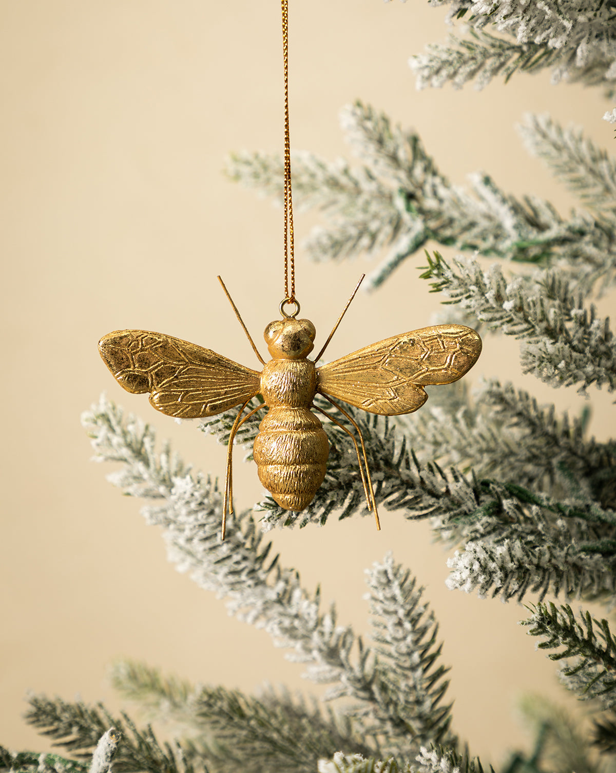 The Golden Bee Ornament, featuring a nature-inspired design, hangs from a gold string on a snow-dusted artificial holiday branch set against a light beige background, by McGee & Co.