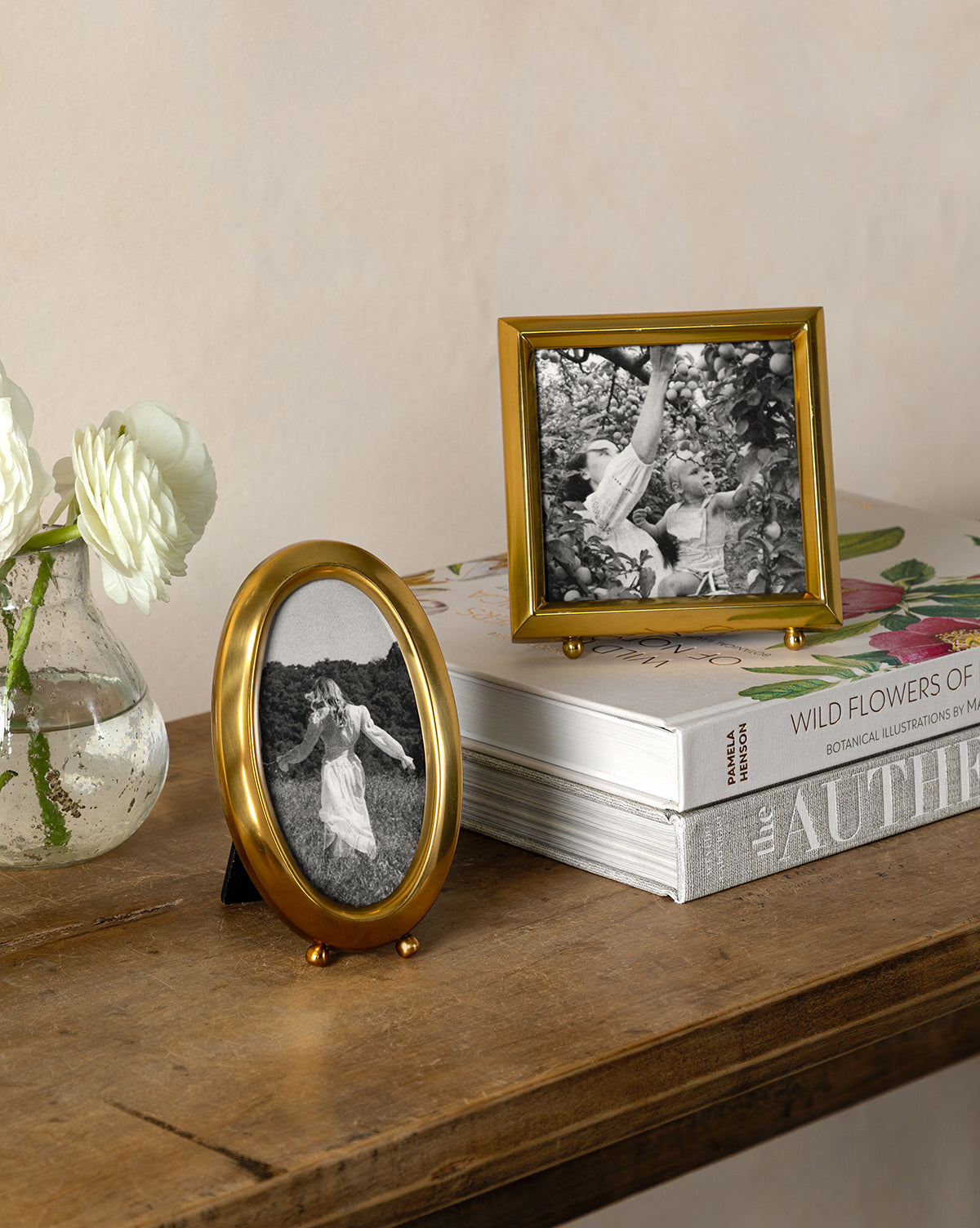 A wooden table holds Wild Flowers of North America, a stack of books, a glass vase with white wildflowers, and two framed black-and-white photos of a girl—one oval, one rectangular.