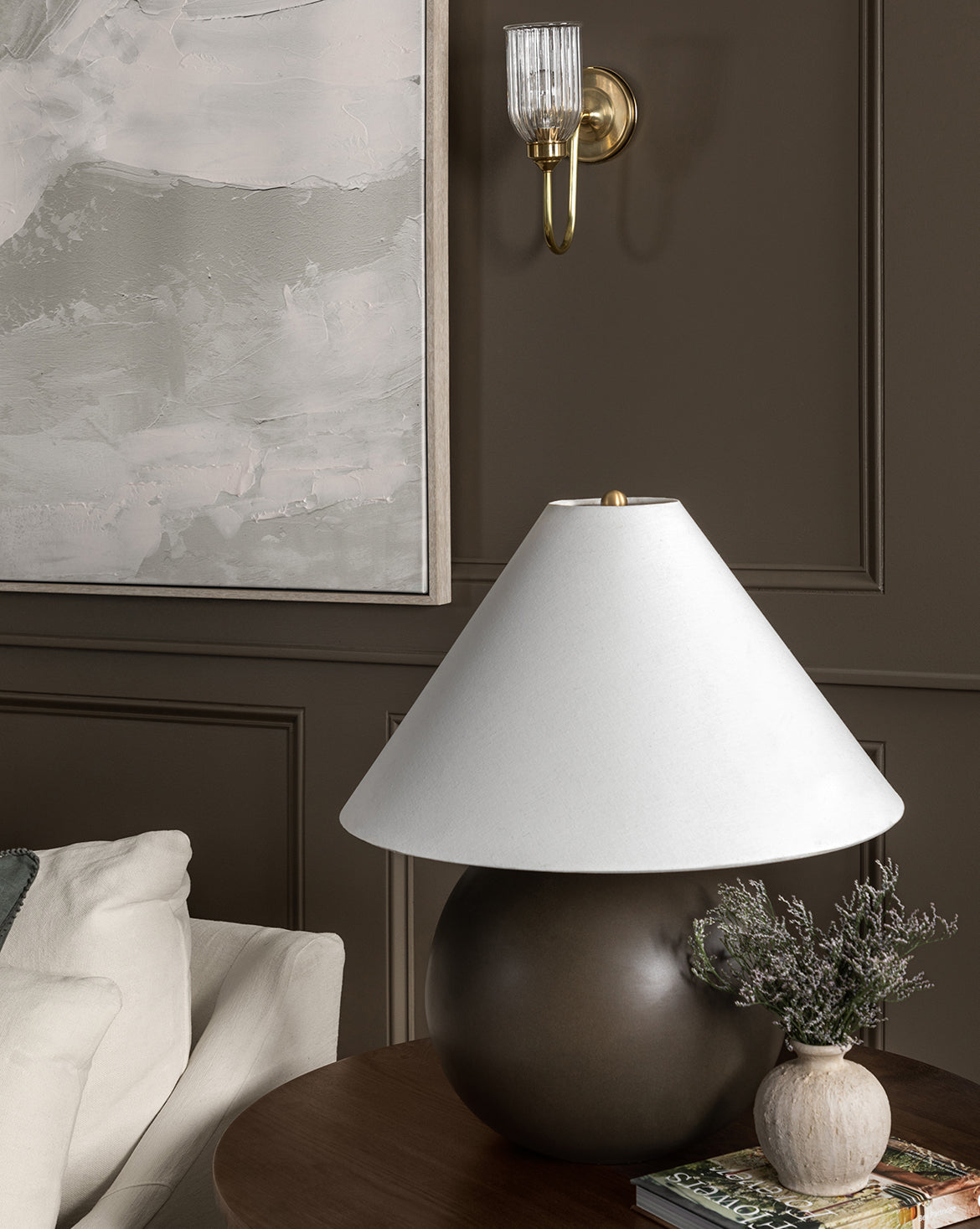 A modern living room features the Tess Large Ceramic Table Lamp with a wide white shade on a wooden table, beside a small lavender vase and books. A cream sofa and abstract wall art are in the background, by McGee & Co.