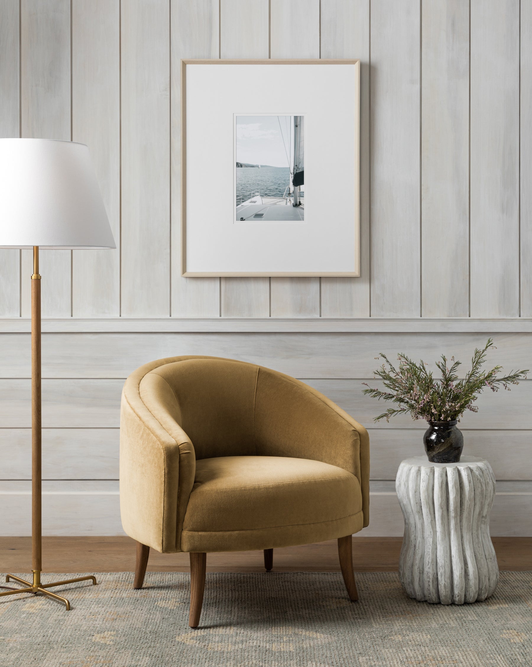 A cozy corner features a light brown velvet armchair, a white floor lamp with a gold stand, a round ribbed side table with greenery, and Sailboat Perspective wall art in a pickled white frame on wood-paneled walls.