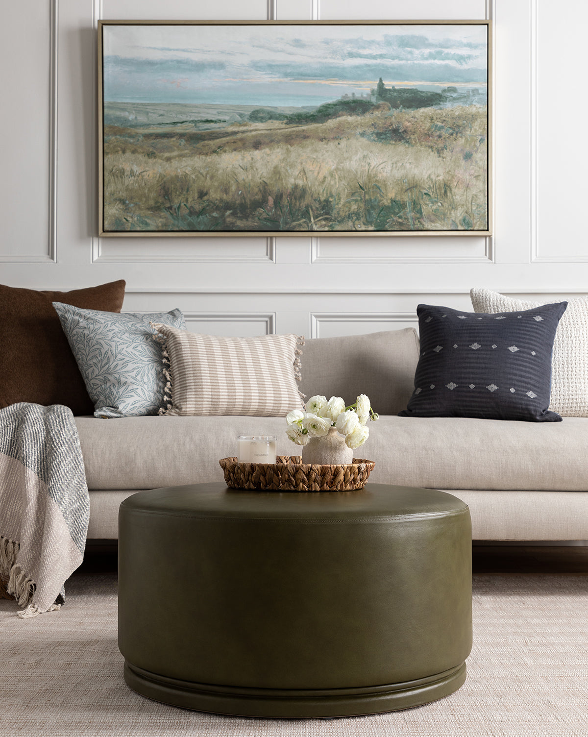 A cozy living room with a beige sofa, patterned pillows, a brown throw, a green round ottoman with flowers and candles, and the Sunset Fields giclee print on canvas in a silver wood frame hanging on the white paneled wall.