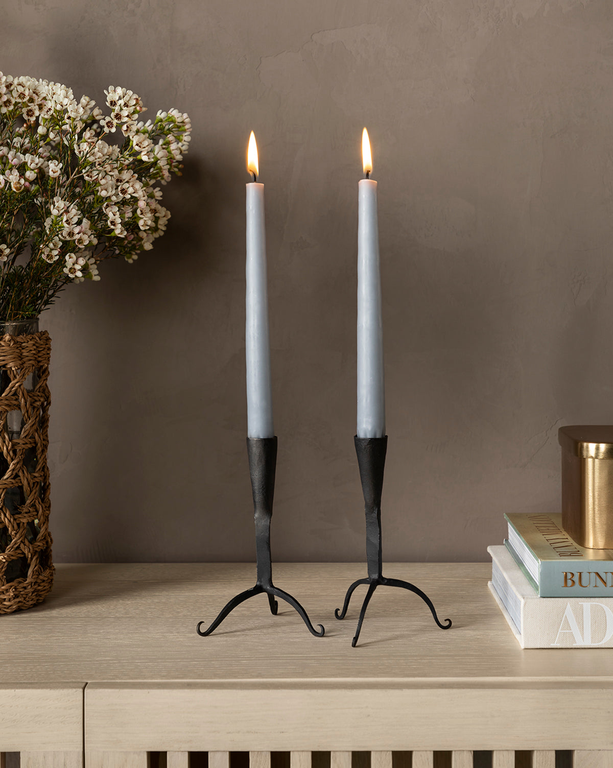 A pair of Light Gray Taper Candles (Set of 12) in black holders casts a warm glow on a light wood surface, next to a woven flower basket and books against a beige wall for a modern touch.