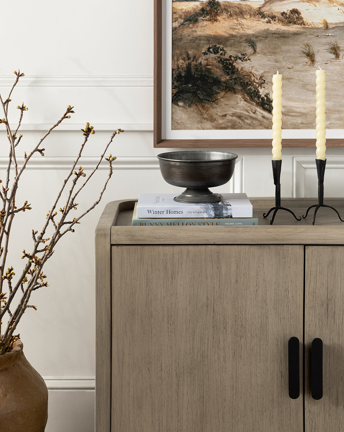 A wooden cabinet with black handles displays a stack of books, the Aged Iron Compote, and two twisted candles in a black holder. Nearby is a vase for flowers, while above hangs a framed landscape painting.