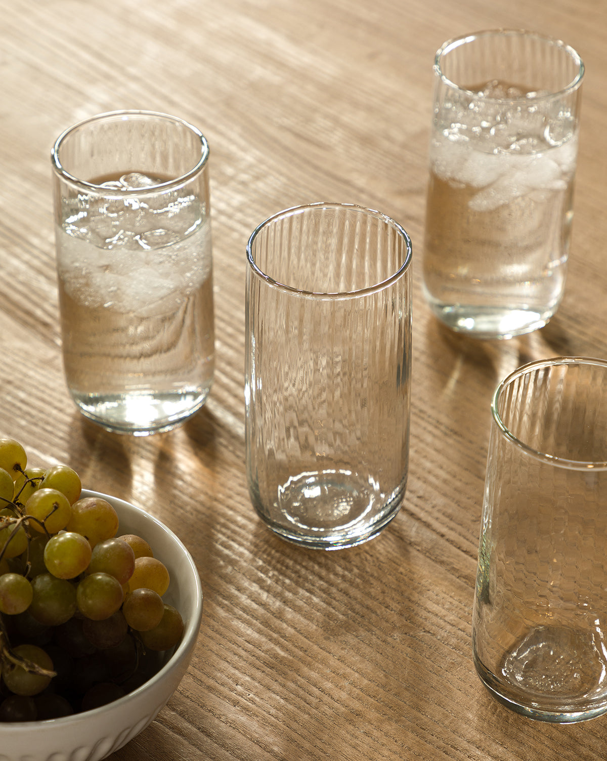 A Ribbed Tall Drinking Glasses (Set of 4) set sits on a wooden table—three filled with ice water and one empty. Their stylish ribbed design is complemented by a bowl of green and red grapes in the lower left corner, by McGee & Co.