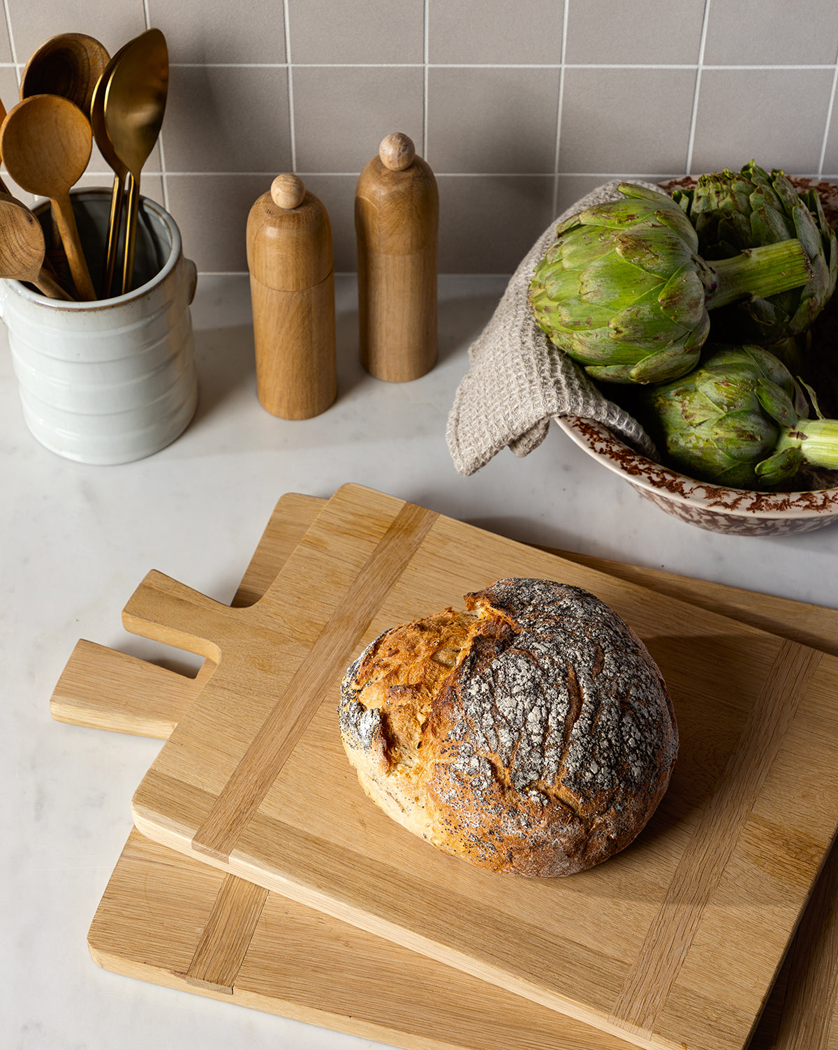 A round loaf of rustic bread sits on the Rectangle Bread Board, made from oak and walnut, stacked on natural-finish cutting boards. Nearby are artichokes, wooden salt and pepper shakers, and a ceramic utensil holder by McGee & Co.