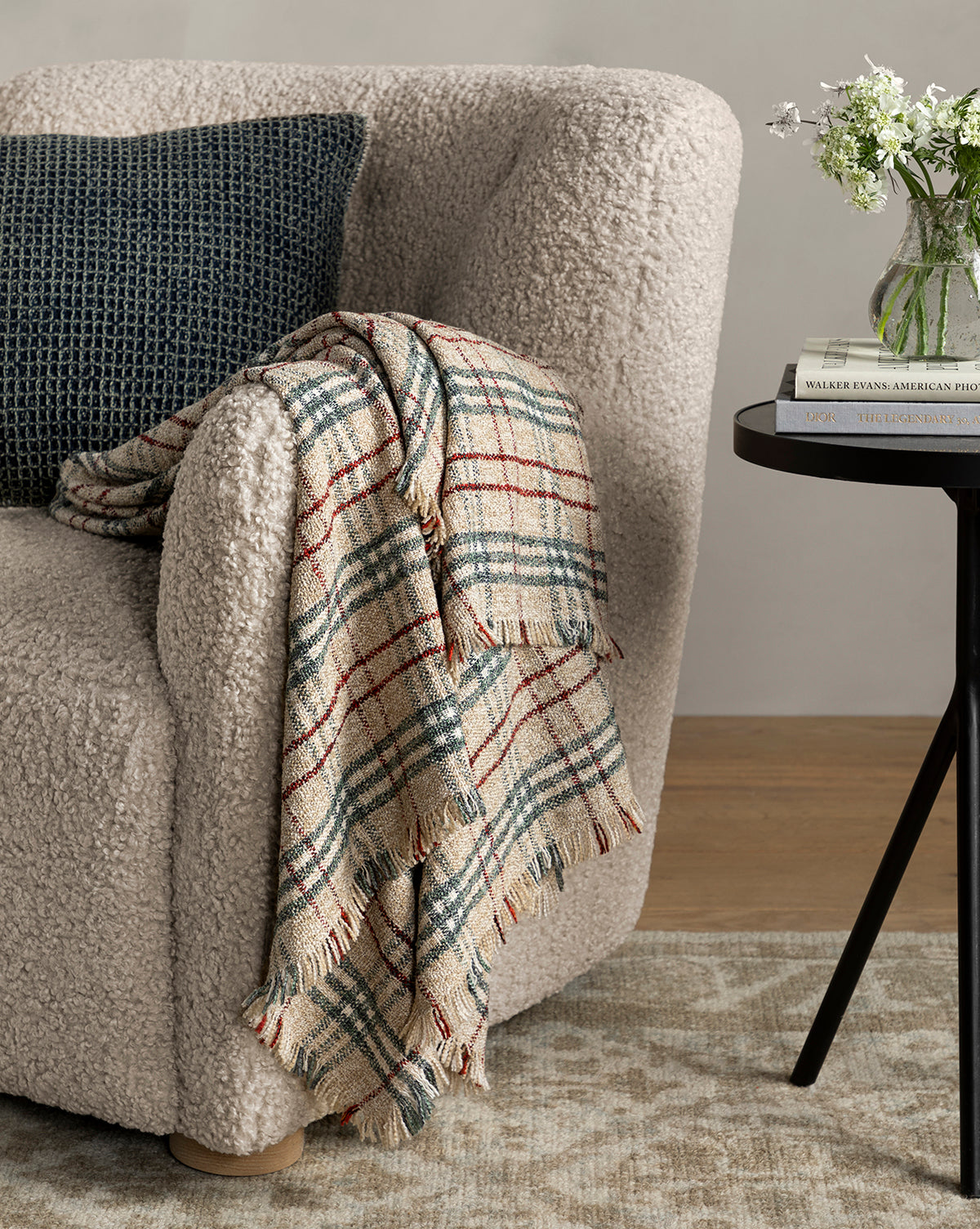 A Magda Lounge Chair (Ready to Ship) with textured upholstery, a dark pillow, and a fringed plaid throw rests beside a round black side table topped with stacked books, a glass vase, and white flowers by McGee & Co.