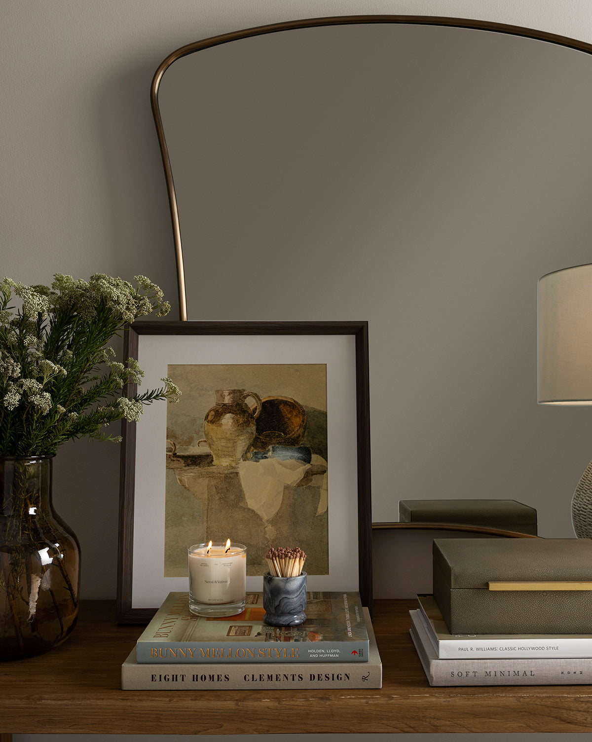 A wooden table holds books, a candle, Tipped Vases with white flowers, a giclee print as vintage wall art, a marble match holder, and a large arched mirror reflecting part of a lamp and the wall.