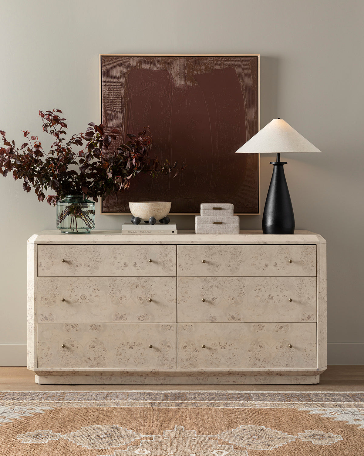 The cream-colored Downtown Brown dresser with six drawers sits against a light wall, topped with dark leafy branches in a vase, a bowl, stacked boxes, and a black lamp. Above it, textural art completes this sophisticated decor scene.