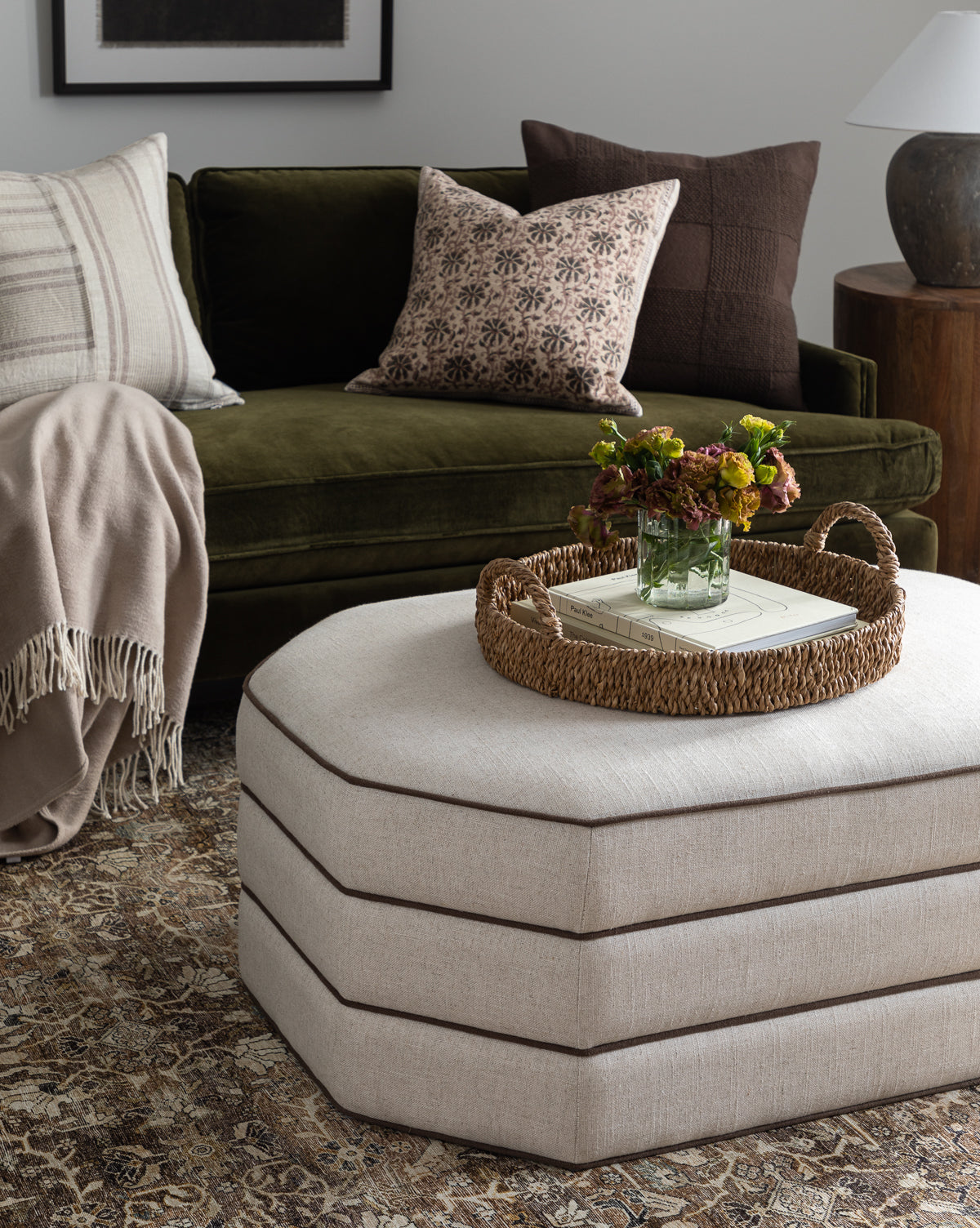 A cozy living room features a green velvet sofa with patterned and solid pillows, a beige blanket, and a light Makerspalm Rafferty Ottoman in performance linen, topped with a woven tray, books, and flowers on a patterned rug from McGee & Co.