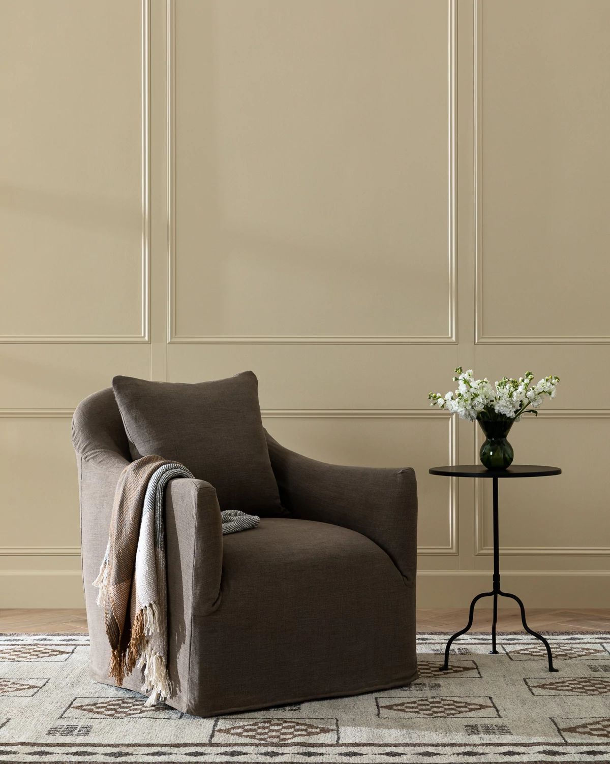 The Trudeaux Slipcover Swivel Chair (Ready to Ship), featuring cozy feather blend cushions, is paired with a pillow and striped throw on a patterned rug beside a round black side table with white flowers against a beige paneled wall by McGee & Co.