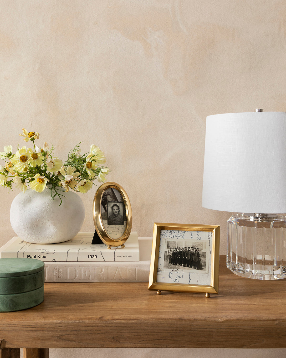 A wooden table displays a white vase with yellow flowers, two Blanche Picture Frames in antique brass, a stack of books, a clear glass lamp with a white shade, and a small green velvet box against a beige wall.